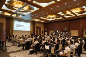 ホテルの煌びやかな一室にて。左奥にプロジェクター、プロジェクターに向かって約100名の参加者が着座して開会の挨拶を聞いている。