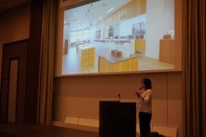 プロジェクターの前に登壇者用の演台が置いてあり、そこで女性の担当者が会社について説明している。
