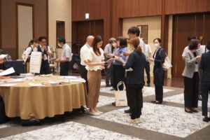 円卓周辺に学校関係者様が立ち留まり、企業様が挨拶に伺っている様子。左サイドに円卓、真ん中に白い服を着た女性（学校関係者）、その学校関係者と懇談しているのは企業様（白シャツの男性と紺スーツの女性）。その周りにも各々懇談している方が数名写っている。盛大な雰囲気が伝わってくる。