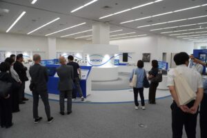 日置電機様の展示スペースにて歴代の製品が展示されている。真ん中にHIOKIの文字パネル、両サイドで男女の見学者が説明を聞いている。