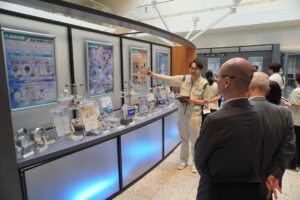 長野計器様の製品が展示されているスペース。作業着を着用した眼鏡をかけた男性が右手で製品上のパネルを指しながら説明している。右サイドに説明を聞いている男性。