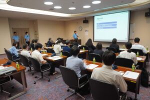 コトヒラ工業様の会議室にて、人事担当者がプロジェクターを使用し会社概要を説明している。プロジェクターの方を向いて約20名の参加者がスクール形式の机に着座し、人事担当者の説明を聞いている。