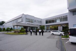 奥に2階建てのユウワ様の自社工場が写っている。正面入り口の方へ、6名の参加者が歩いて行く写真。